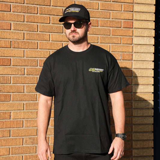 Man wearing a black t-shirt with a logo and a cap, standing against a brick wall.