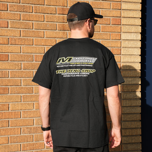 Person wearing a black t-shirt with a logo and text, standing against a brick wall.
